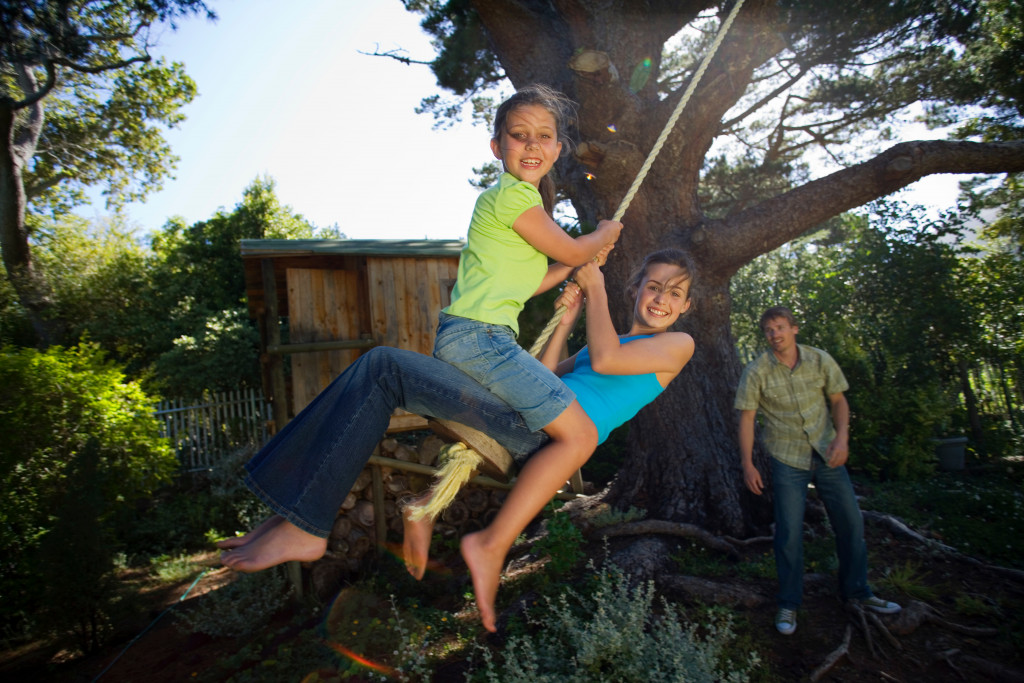 Portrait of two young girls swinging on a garden rope swing near the treehouse with father pushing in the background.