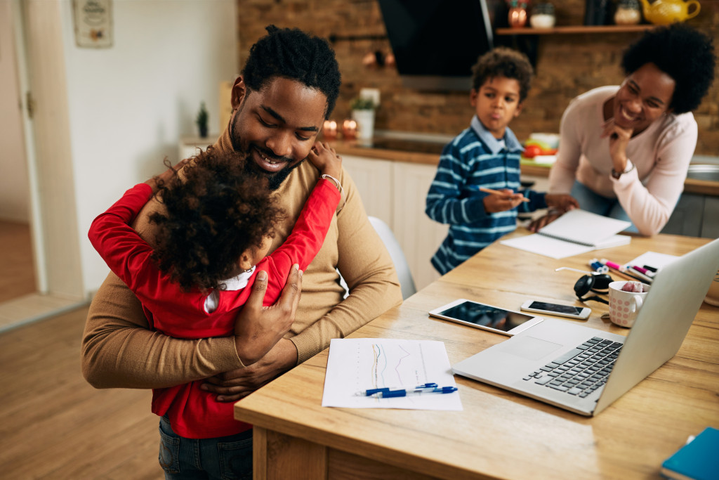 Happy African American man embracing his small daughter while working on a computer at home. Mother and son are in the background.