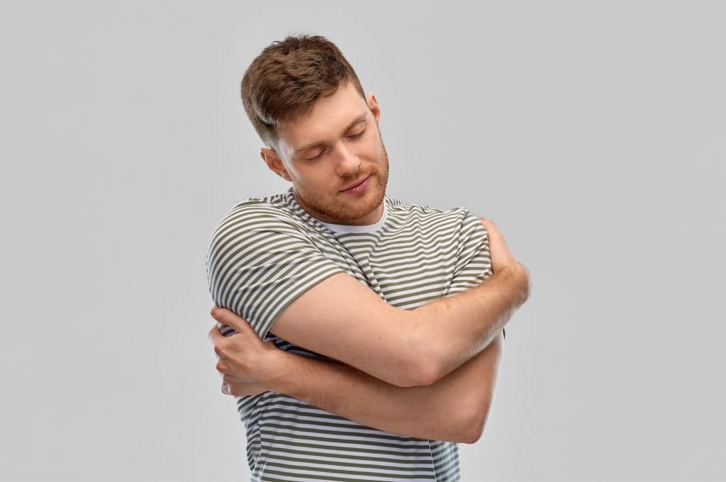 happy young man in striped t-shirt hugging himself over grey background