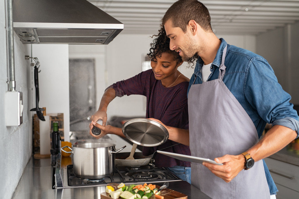 Happy girl adding salt in pot for pasta while guy hold the lid up